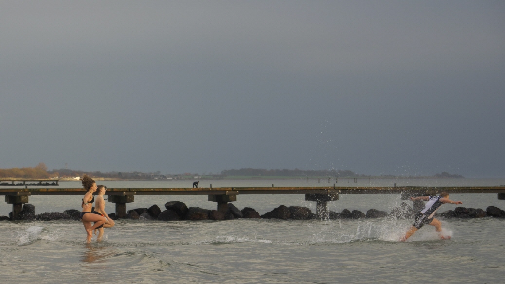 Menschen rennen in die Ostsee und die langen Haare wehen im Wind. Mann mit Fußballtshirt spritzt mit Wasser